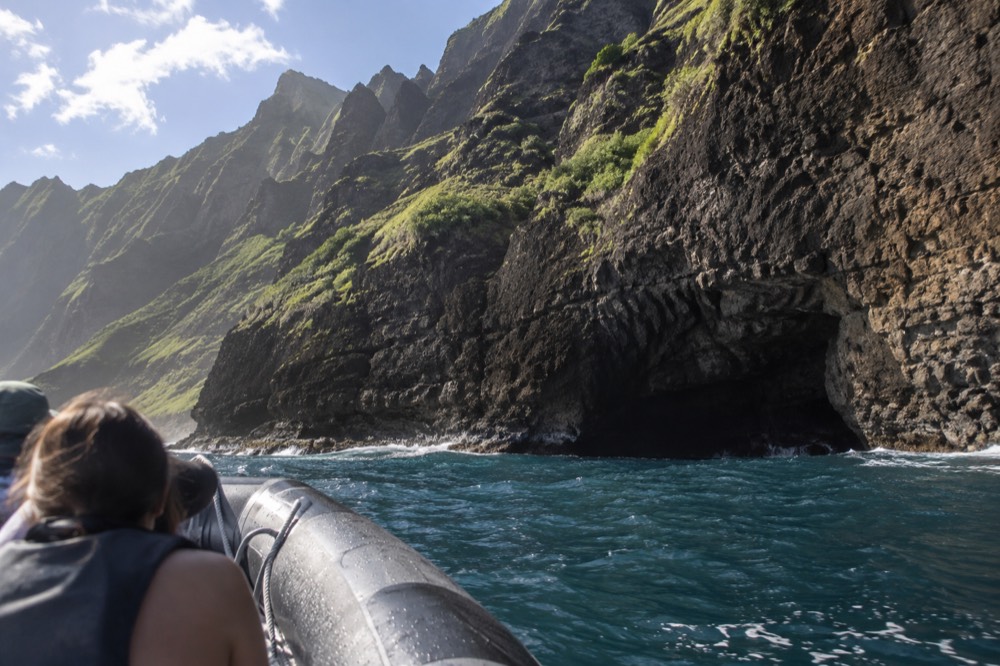 Small inflatable raft cruising close to a tall sea cliff with a dark cave opening at the waterline