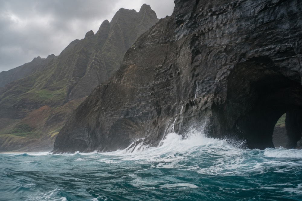 Ocean waves breaking against the base of a steep coastal cliff near a dark rock opening