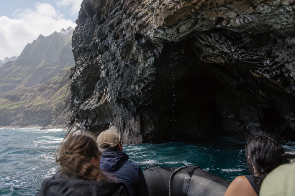 A rigid inflatable raft cruising close to a dark sea cave entrance in a cliff face.