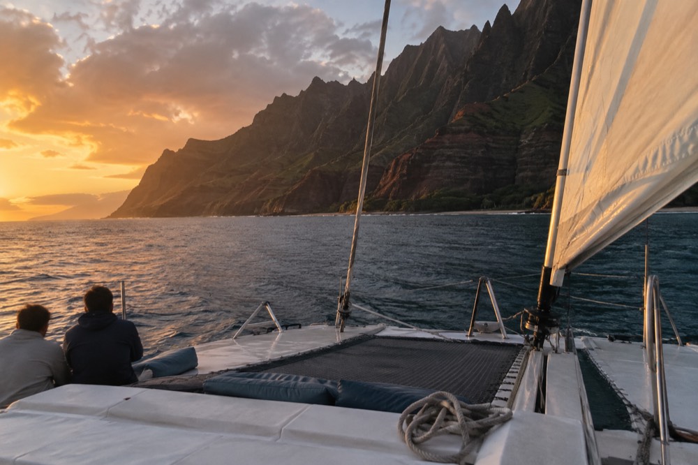 A catamaran under sail on calm evening water near a rugged island shoreline