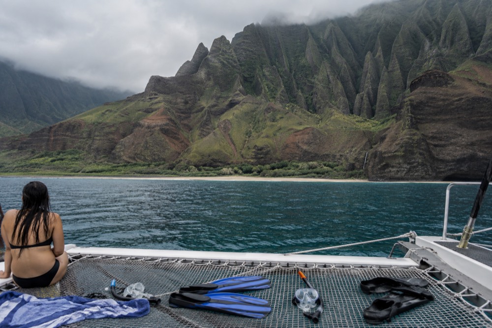 A catamaran at anchor in a quiet cove beneath green Napali cliffs, with snorkel gear laid on the deck.