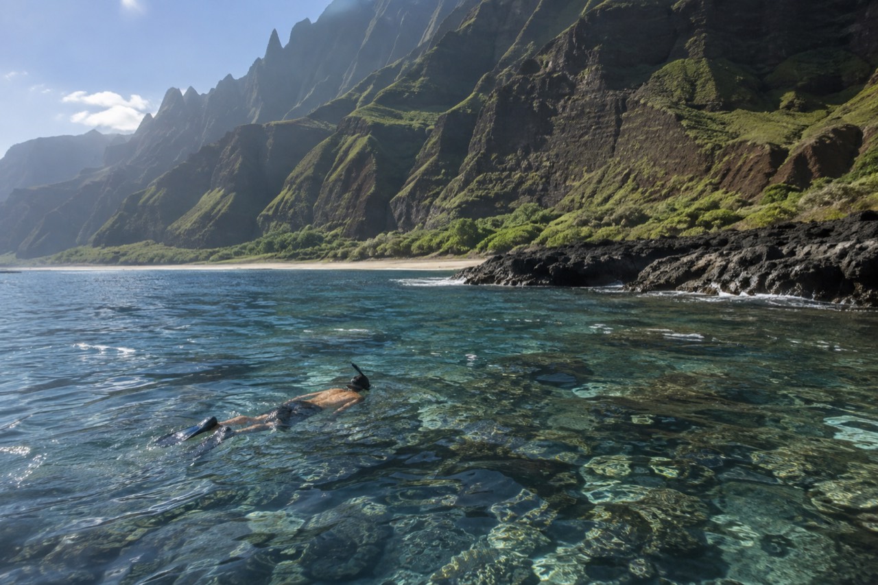 A snorkeler floating above a coral reef beside the cliffs of the Napali Coast on Kauai.