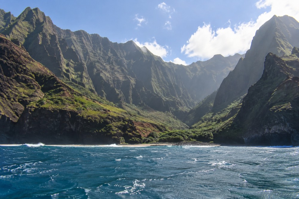 Sunlit Napali Coast cliffs and green valleys under a bright midday sky seen from the water