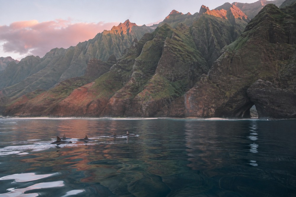 Towering emerald cliffs of the Napali Coast reflected in calm ocean water with a sea arch in the distance