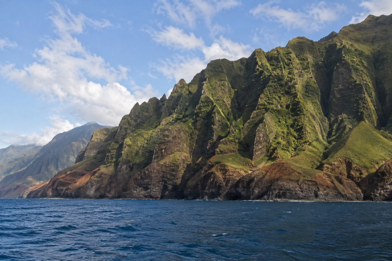 Napali Coast sea cliffs rising from calm blue water with green ridges under scattered clouds