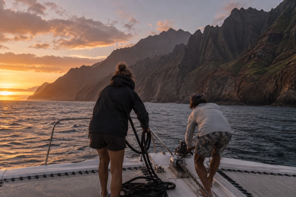 Two crew members coiling rope on the bow of a boat in evening light