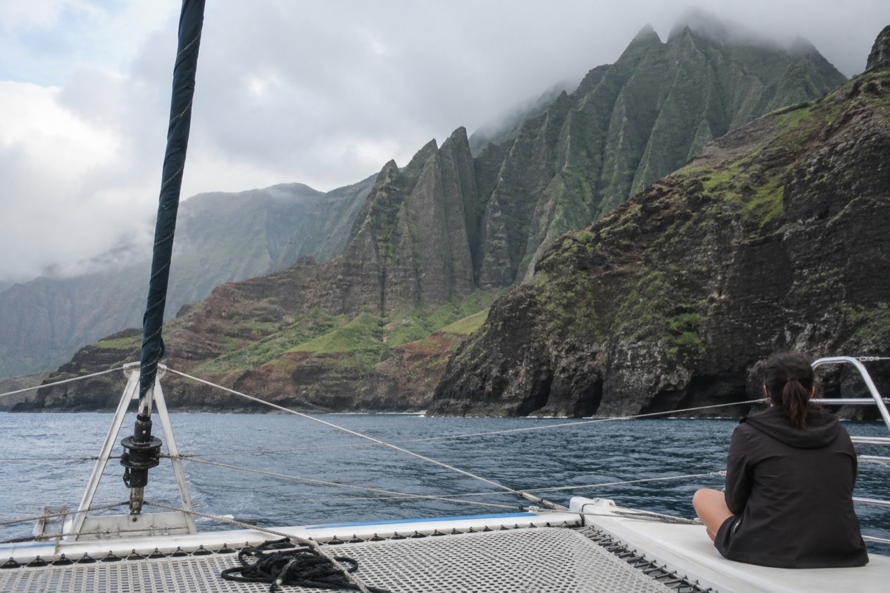 A catamaran sailing along the green cliffs of the Napali Coast on a calm morning