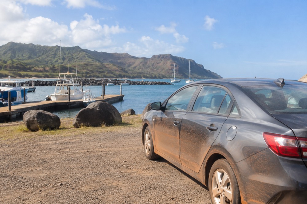 A parked rental car beside a small boat harbor with tour boats at their moorings