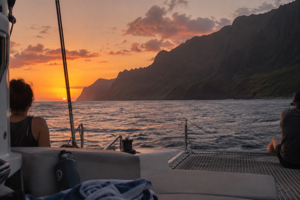 A sailing catamaran anchored near sea cliffs as the sun sets over the Pacific