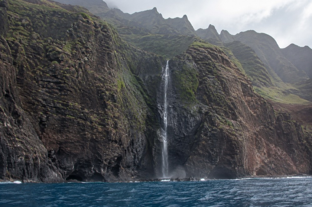 Tall green cliffs and a waterfall along the Napali Coast seen from open water