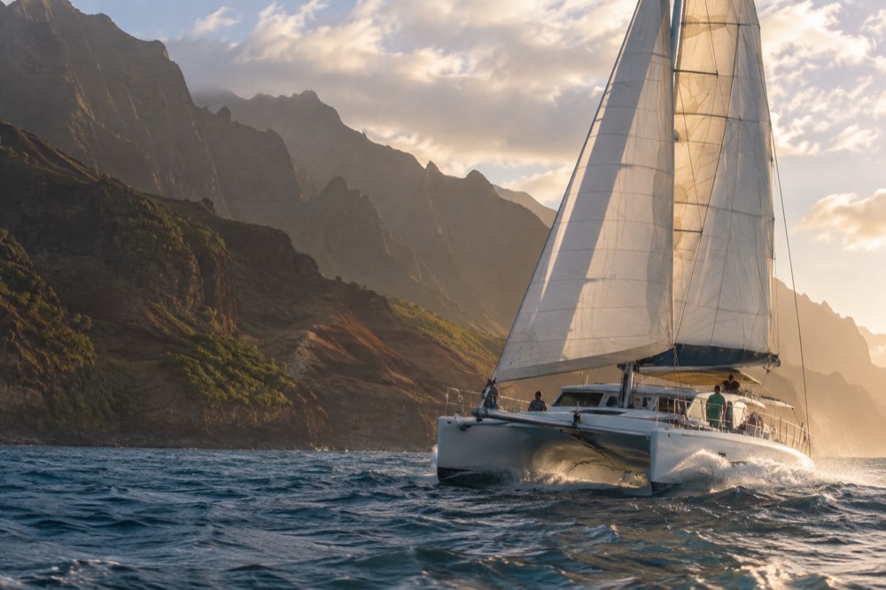A catamaran under sail near sea cliffs on the Napali Coast in late afternoon light