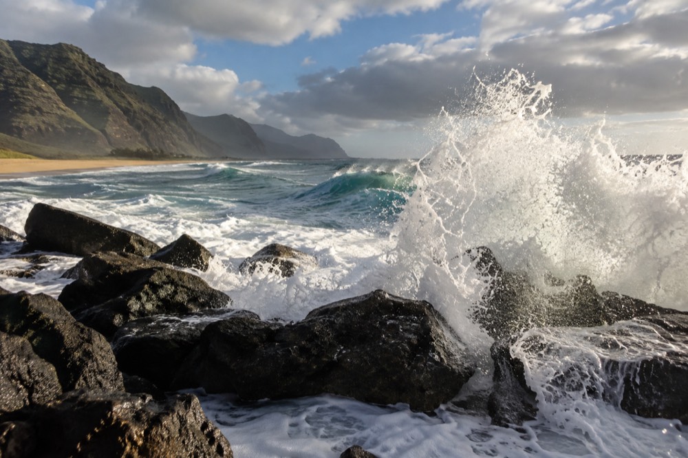 Deep blue ocean swell breaking against a rocky Hawaiian shoreline under a partly cloudy sky