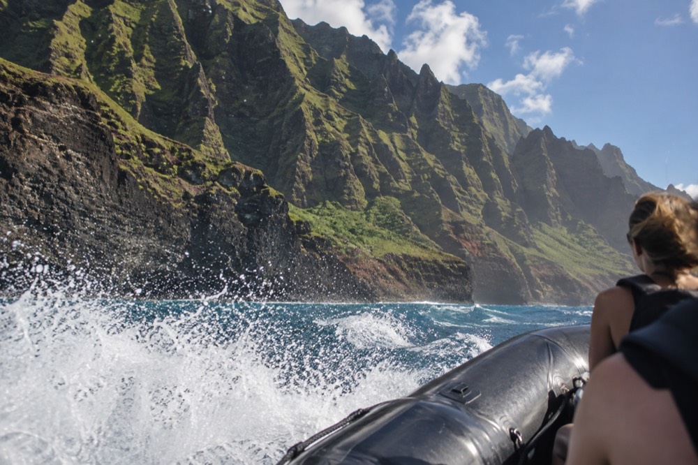 A rigid inflatable raft with spray flying over the bow tubes along a steep green cliff face