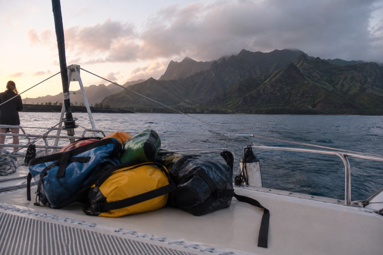 A catamaran deck with dry bags and jackets stacked beside the railing as cliffs rise in the distance