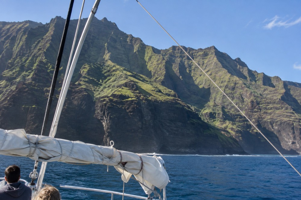 A catamaran under sail passing the green ridged cliffs of the Napali Coast in clear morning light.