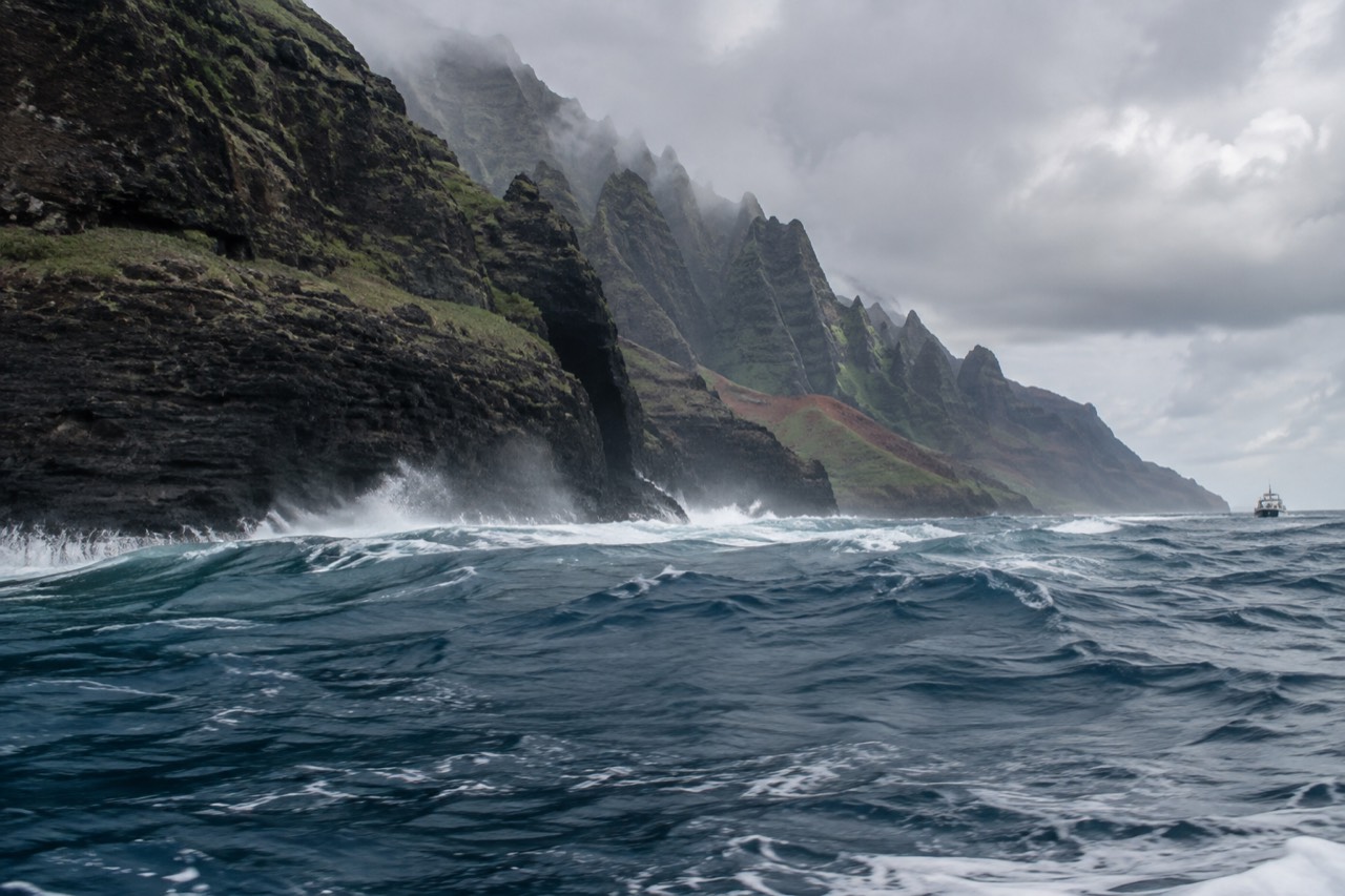 Dark ocean waves breaking against the steep green cliffs of the Napali Coast under a grey winter sky.