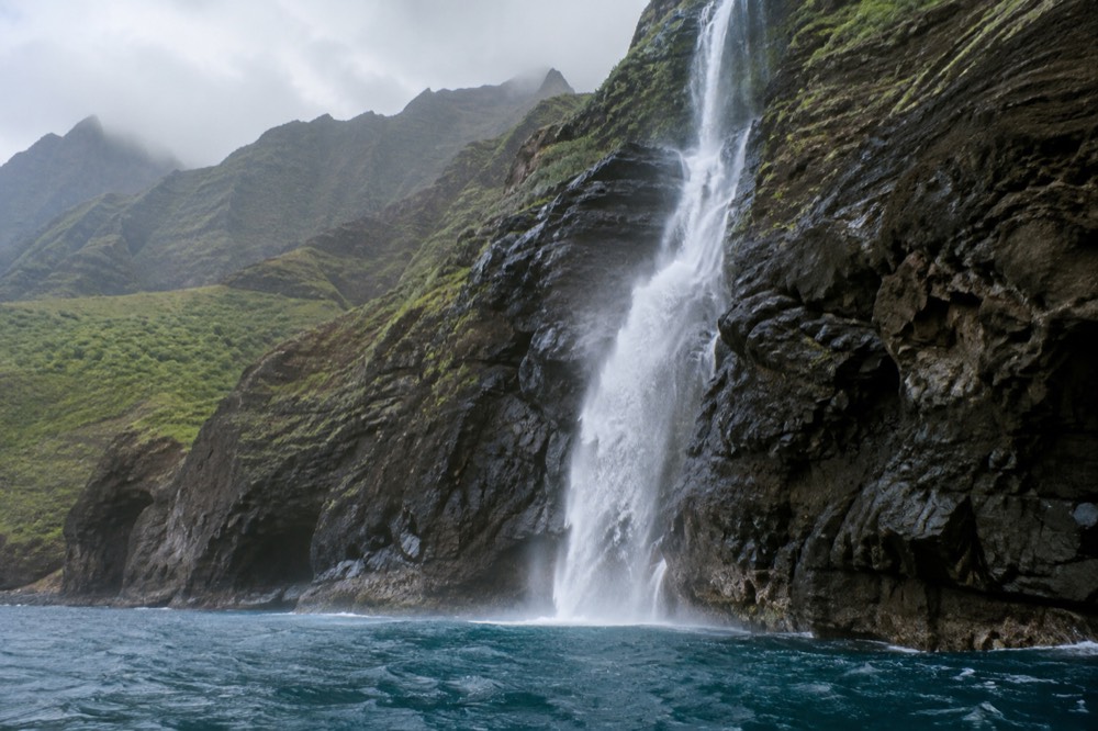 Napali Coast cliffs with a waterfall cascading into the ocean and a small boat in the foreground