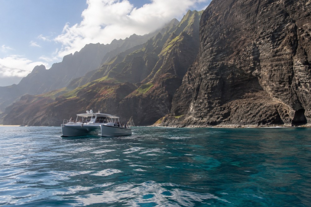 A wide catamaran anchored near a tall sea cliff with clear water below