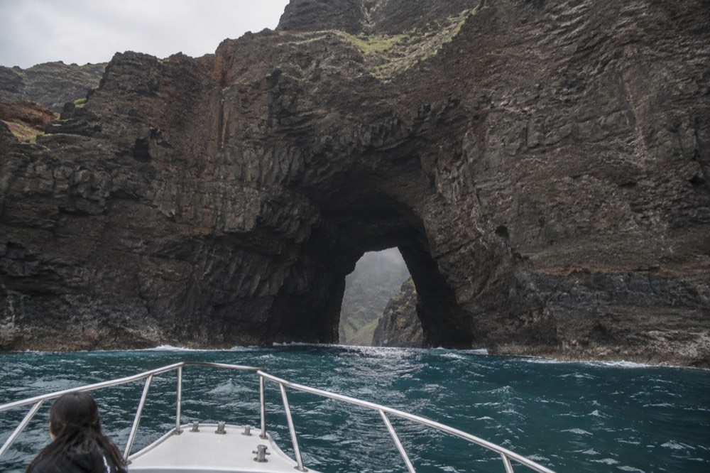 A tour boat approaching a tall sea cliff with a narrow valley opening along the Napali Coast