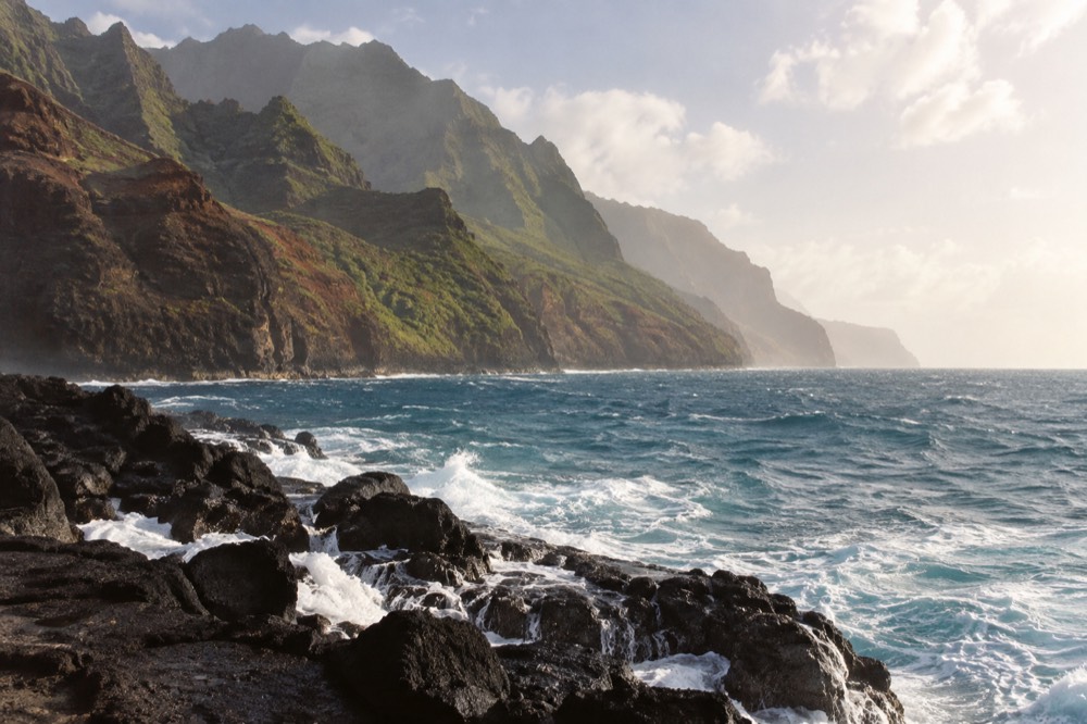 Open ocean swell rolling toward the green cliffs of the Napali Coast under a bright sky.