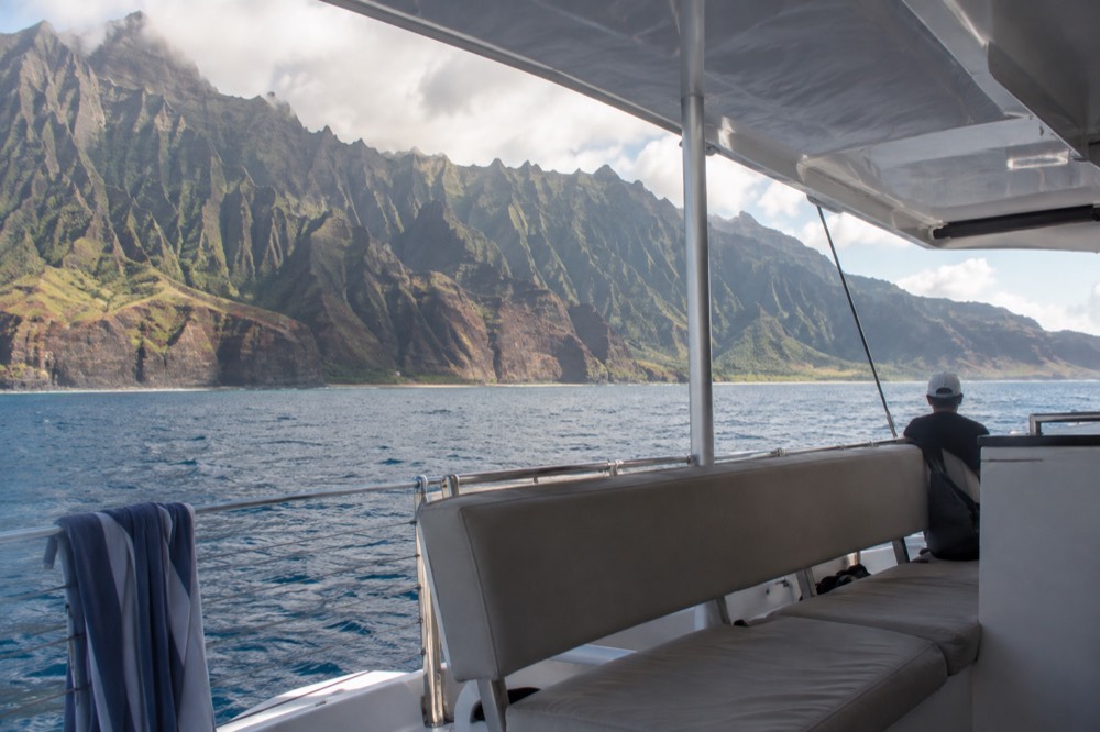 Passenger seating along the mid-deck of a catamaran, facing the Napali cliffs across calm water.