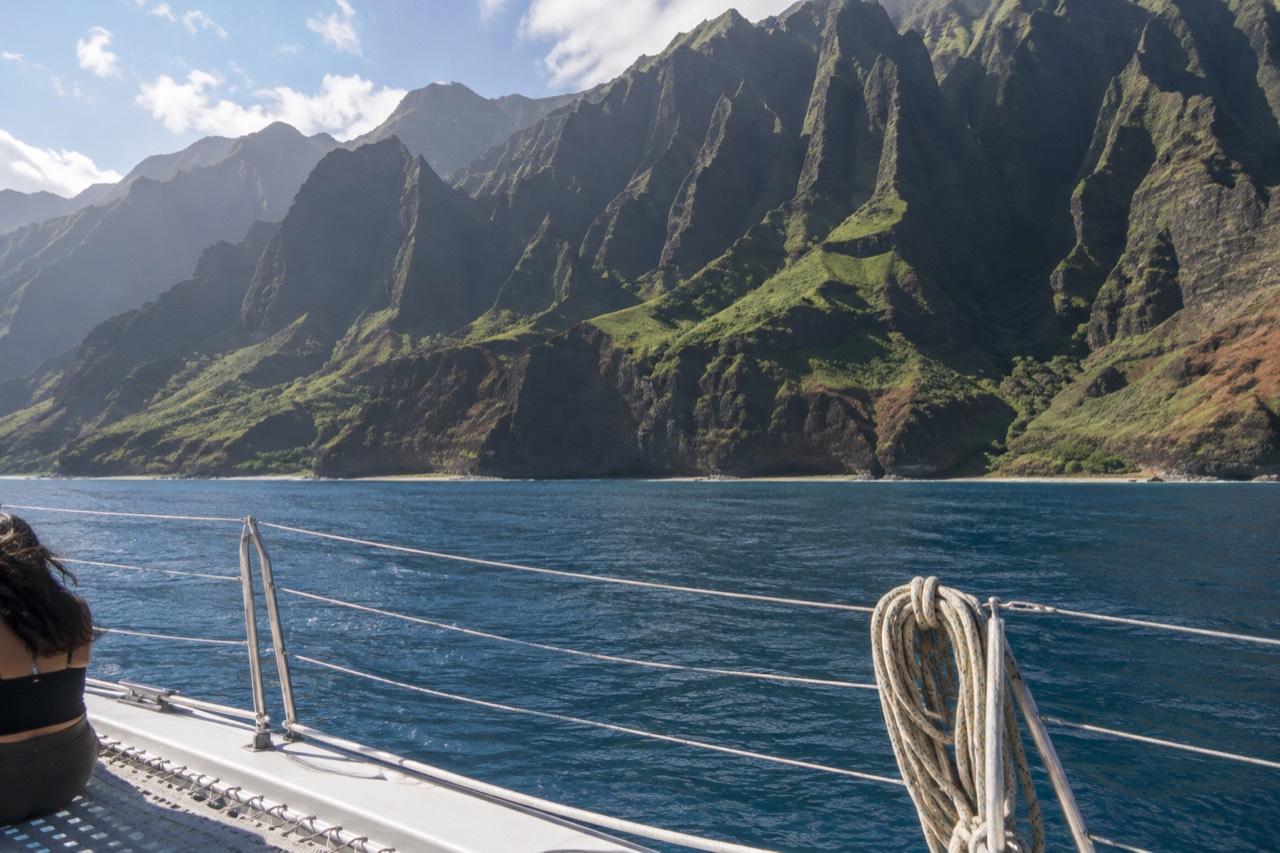 A catamaran cruising along the sheer green cliffs of the Napali Coast on calm blue water.