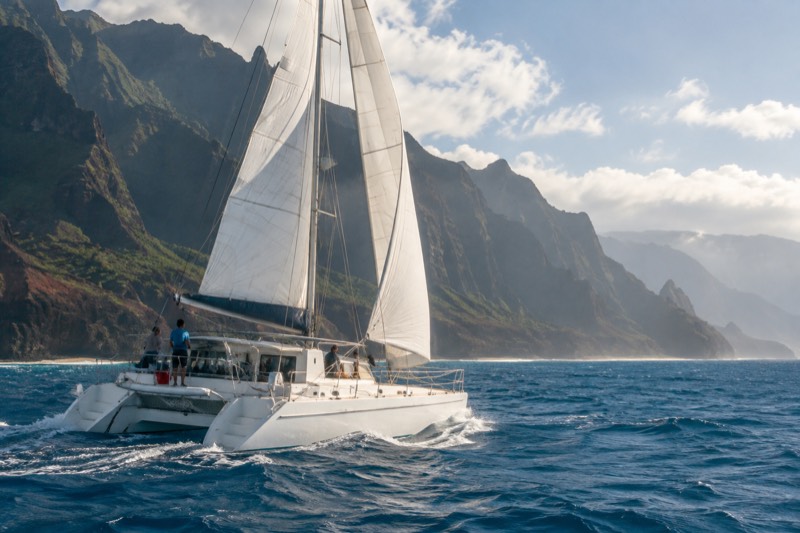 A sailing catamaran heeling under raised white sails on open water off the Napali Coast of Kauai, the three sail-powered trips compared in this section.