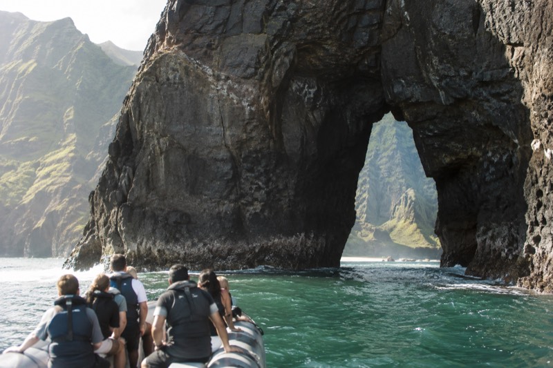 A rigid-hull inflatable raft running close under the Napali sea cliffs on Kauai beside a dark sea arch, the trip style behind the six raft tours compared in this section.