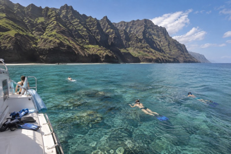 Snorkelers in clear turquoise water beside a catamaran anchored over a Kauai reef, the in-water stop that defines the six snorkel cruises compared in this section.