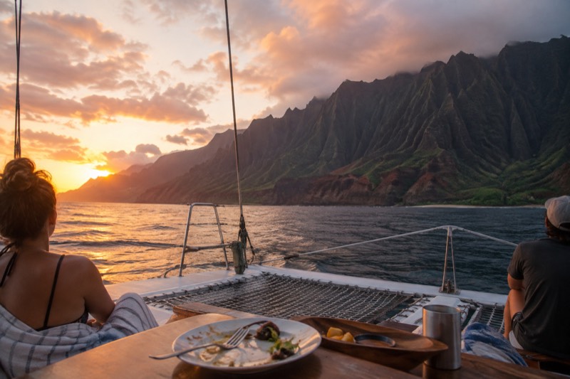A catamaran on gold evening water below the Napali cliffs on Kauai, the setting for the nine sunset and dinner cruises compared in this section.