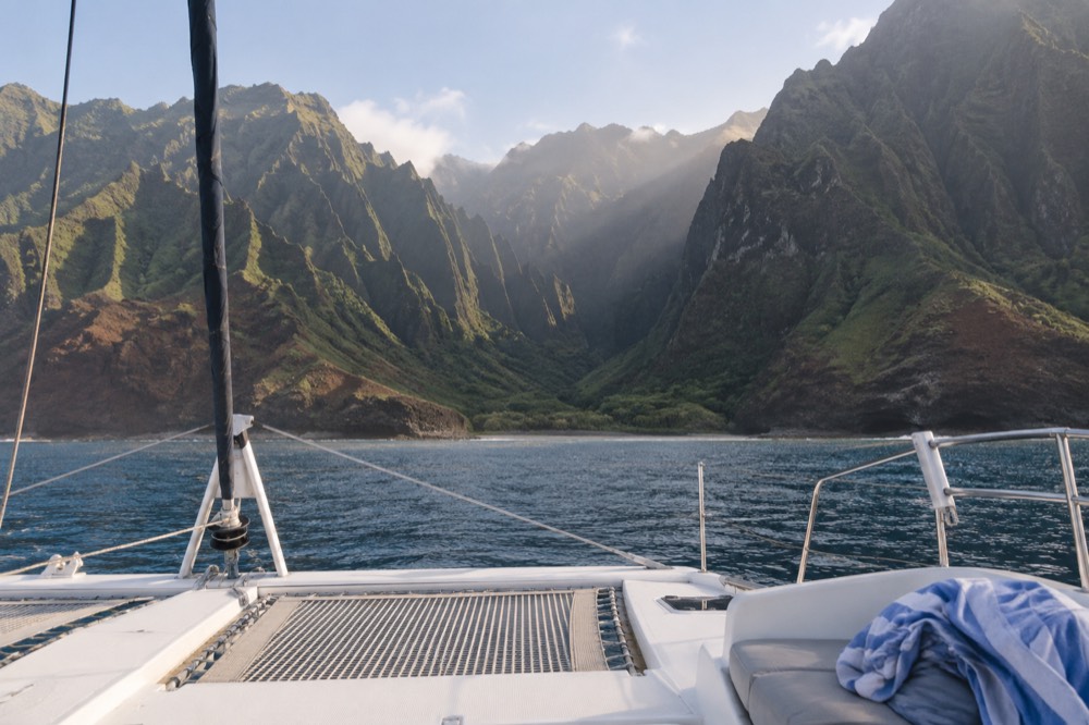 A sailing catamaran anchored near tall coastal cliffs with green ridges above