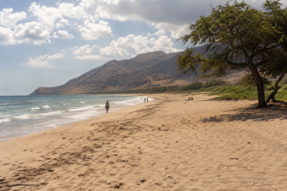 A wide sandy beach with gentle waves and mountains in the distance under scattered clouds