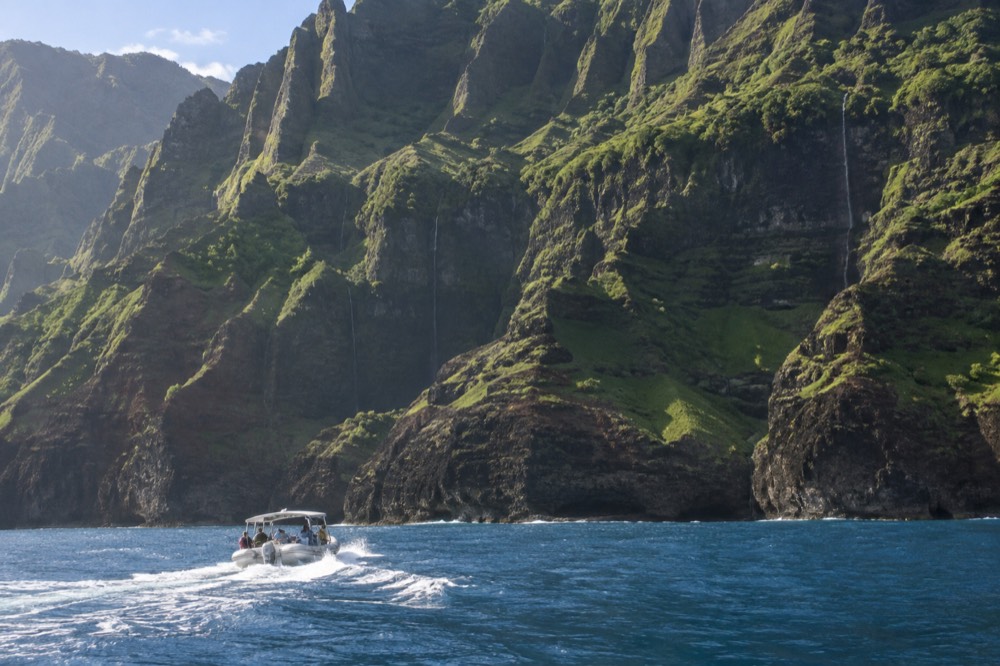 A boat underway near tall green sea cliffs with waterfalls streaking the rock face