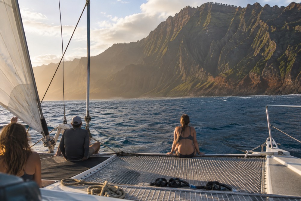 A sailing catamaran heels gently under canvas on long afternoon swells beneath the fluted basalt cliffs of the