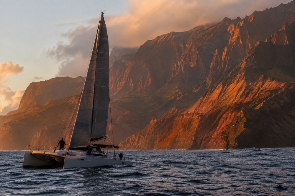 A catamaran under sail on open water with cliffs glowing orange in evening light
