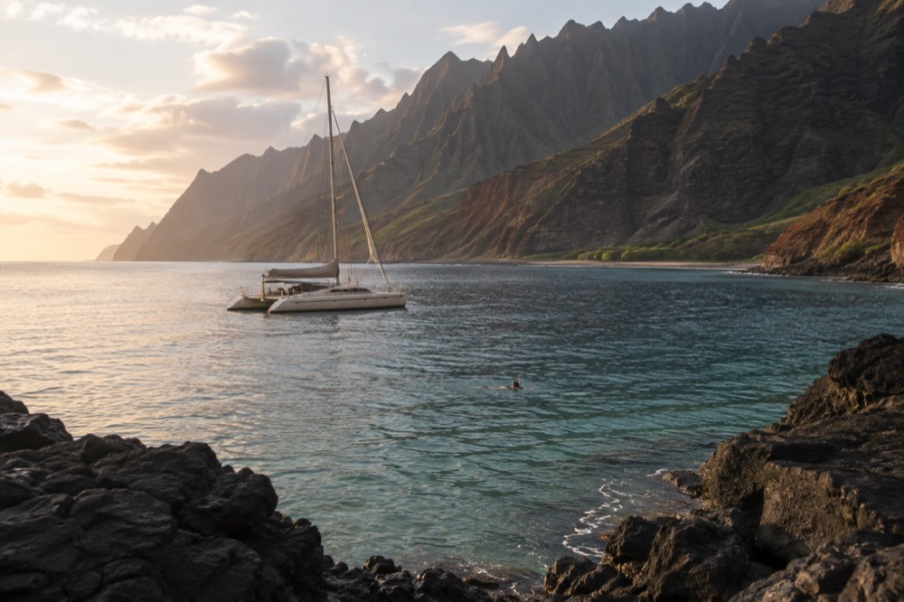 A sailing catamaran at anchor in a quiet cove with warm evening light on the water