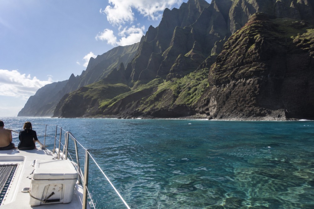 A catamaran cruising close to green Napali cliffs above clear water