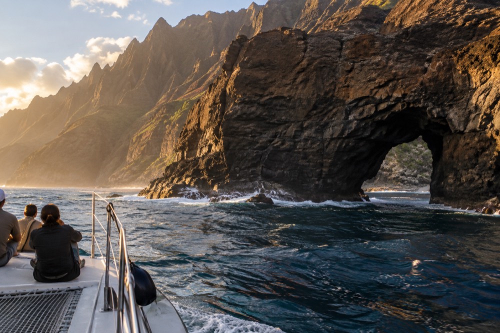 A catamaran under way along the Napali Coast near a sea arch in golden light