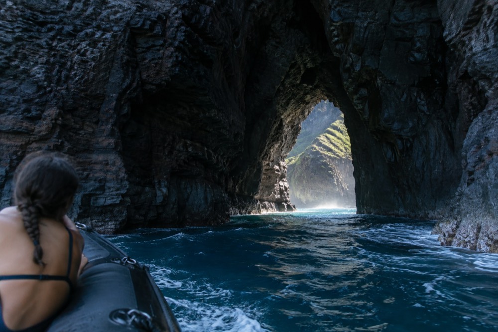 A rigid-hull raft idling at the mouth of a shadowed sea-cave opening near Honopu Arch