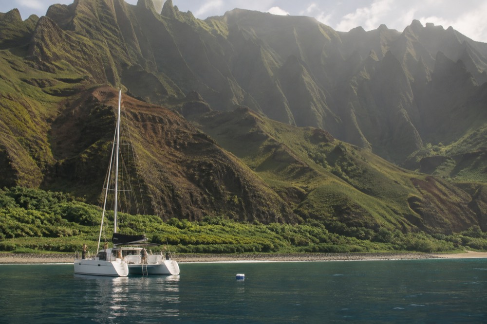 A twin hulled catamaran anchored near the base of tall sea cliffs with green ridges behind