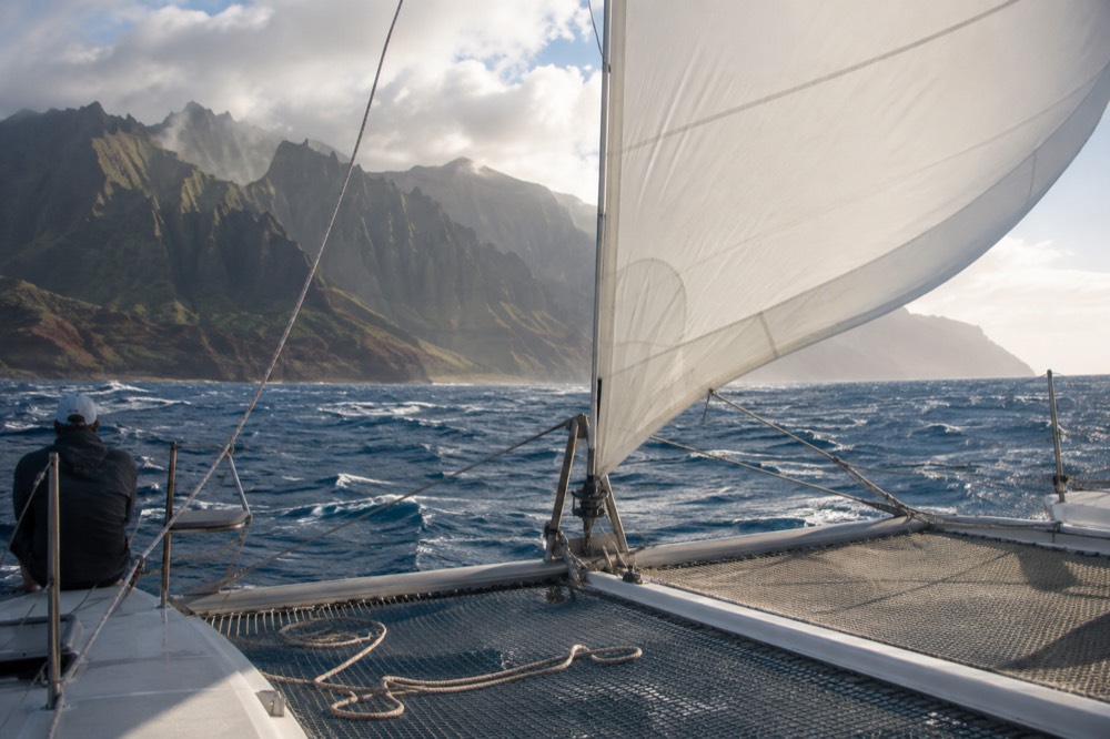 A sailing catamaran under full sail on open water with distant coastal ridges