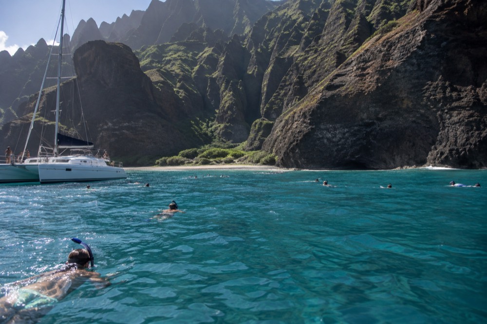 A catamaran at anchor in a quiet cove with snorkelers in the water near the cliffs