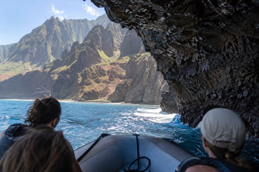 A low rigid-hull raft nosing under a wave-cut overhang at the waterline on Kauai
