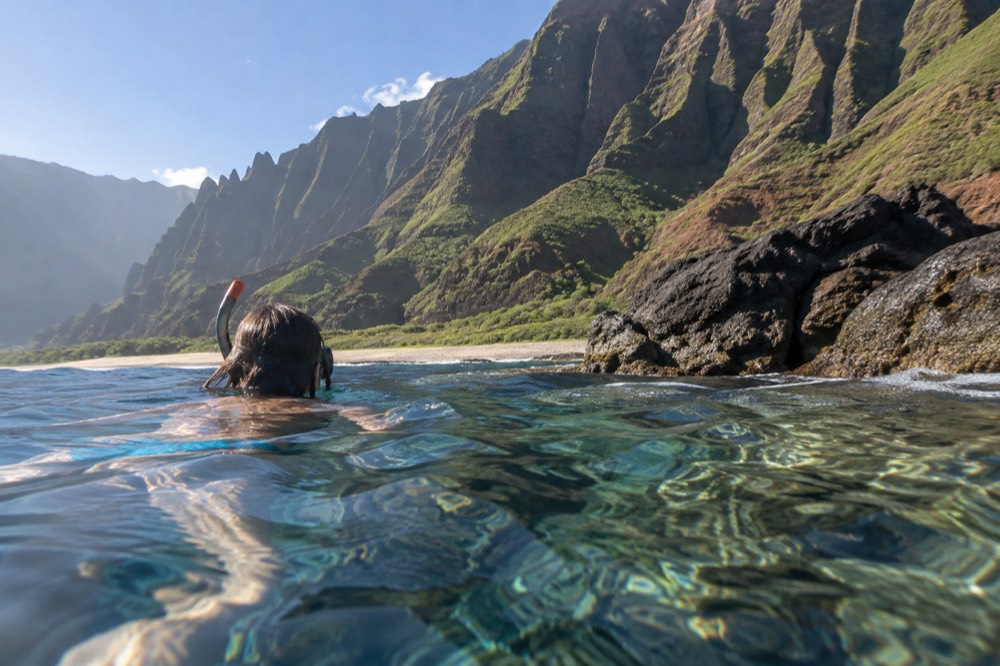 A snorkeler floating over a shallow coral reef in clear water near dark rock