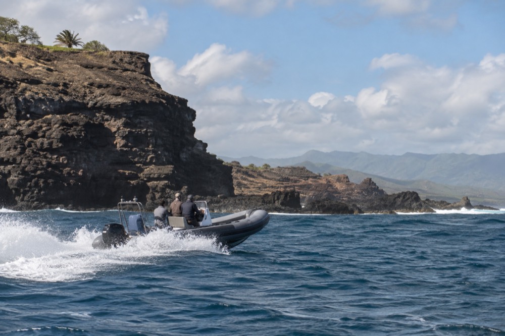 A rigid-hull inflatable raft cruising near a rocky shoreline with sea cliffs behind