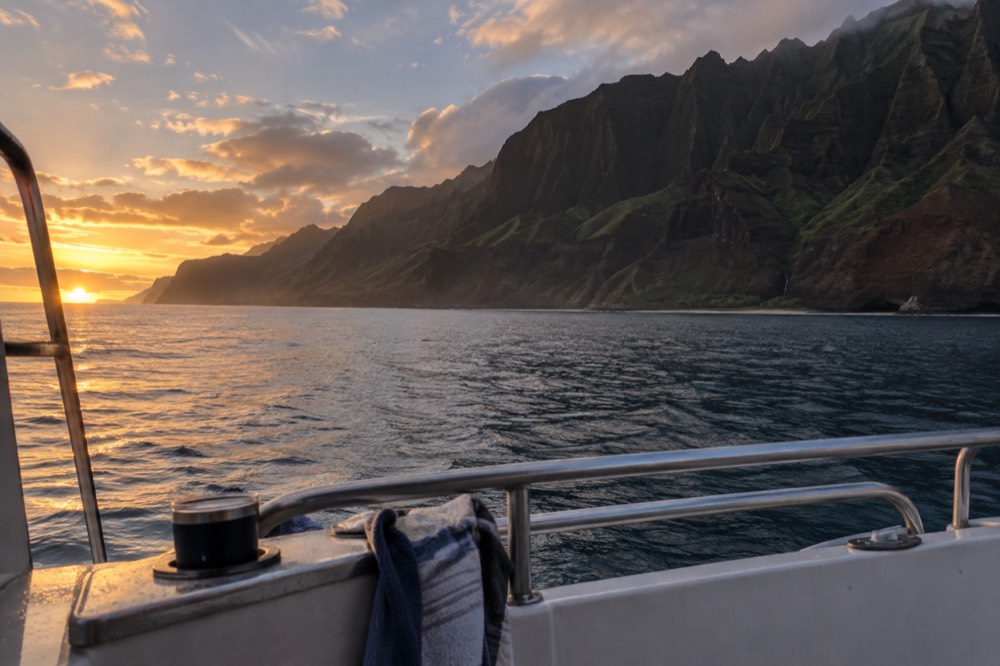 A boat deck at sunset with orange sky over calm ocean and dark cliffs