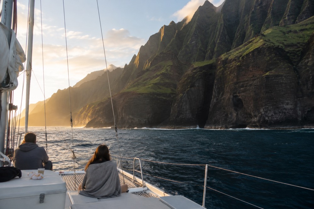 A catamaran cruising close to tall green sea cliffs along a tropical coastline