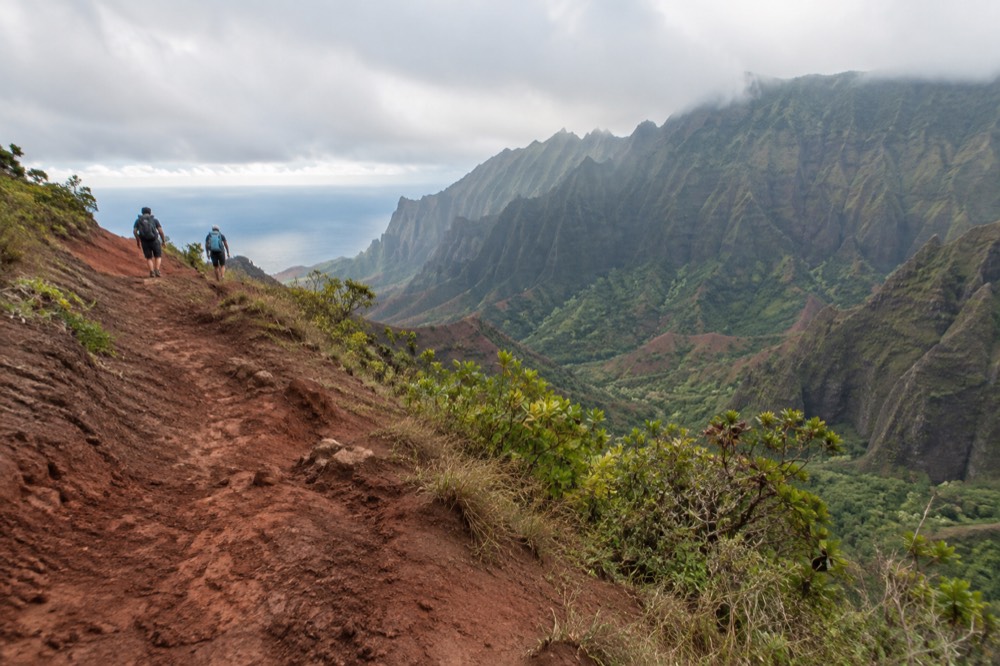 Hikers on a red dirt trail with the Waimea Canyon stretching out behind them